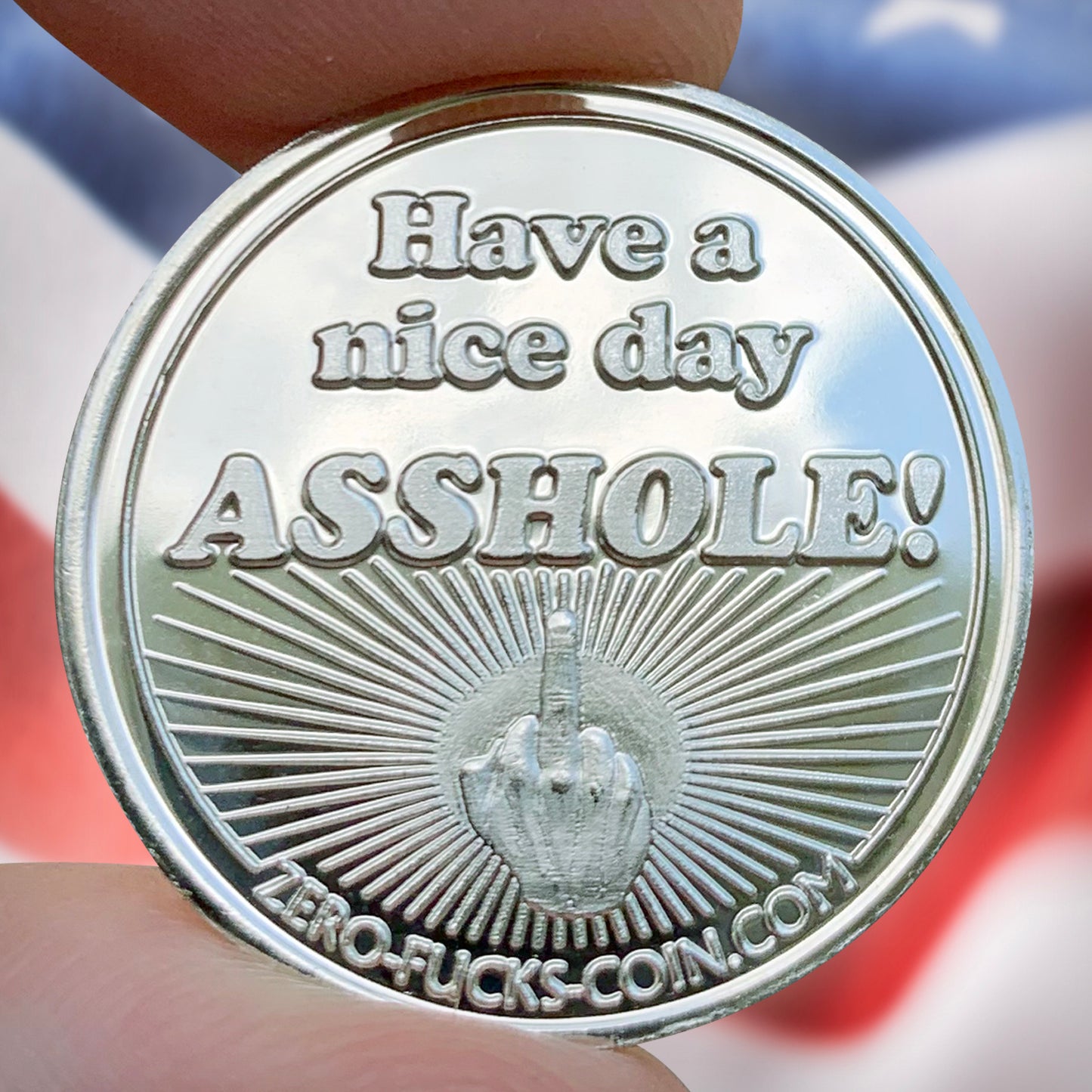 Silver coin with text 'Have a nice day ASSHOLE!' held by a hand against an American flag background.