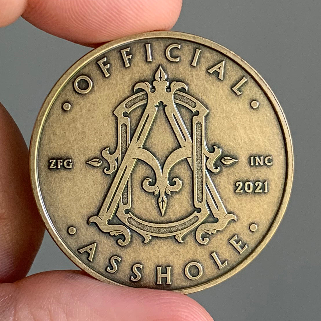 Bronze coin with 'OFFICIAL ASSHOLE' text and design held by a hand against a gray background