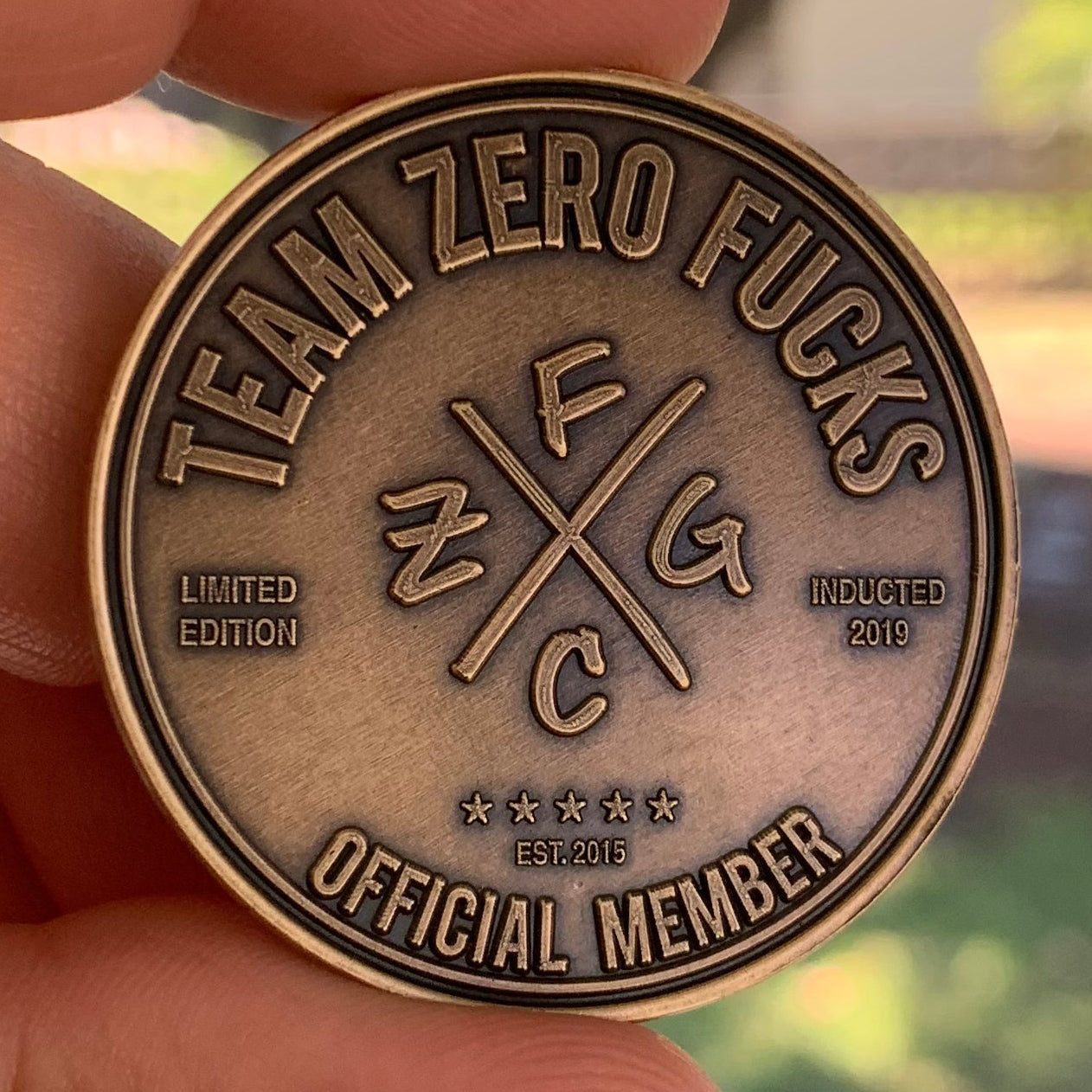Bronze coin with 'Team Zero Fucks' text held in a hand outdoors.